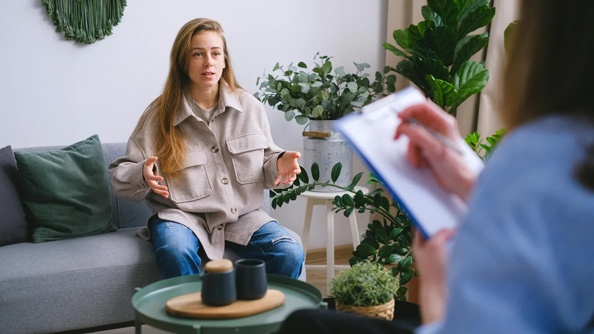 A person on a couch in conversation with a therapist, warm room with plants.