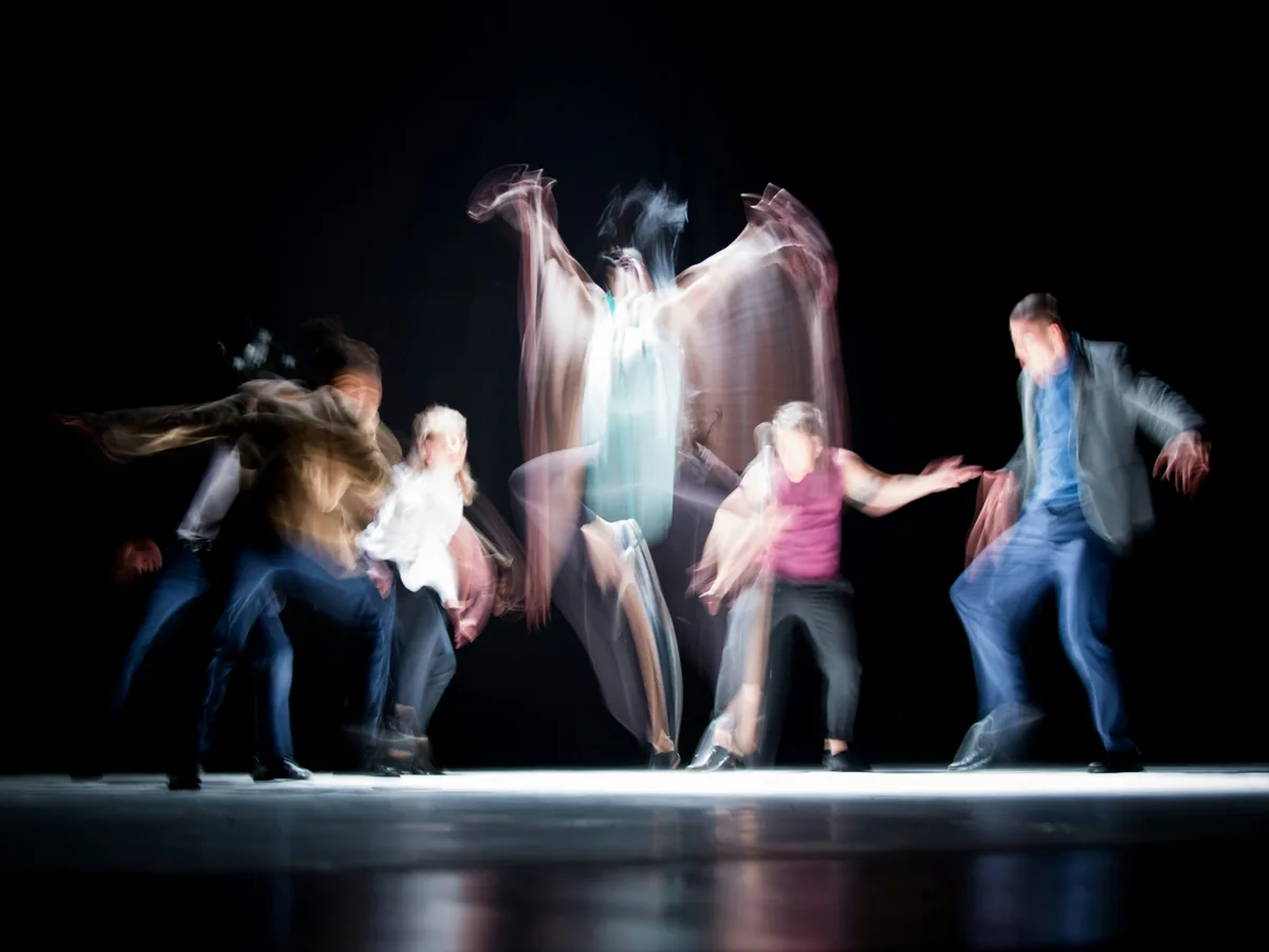Performers in motion on a darkened stage, captured in a long exposure that traces movement and gesture.